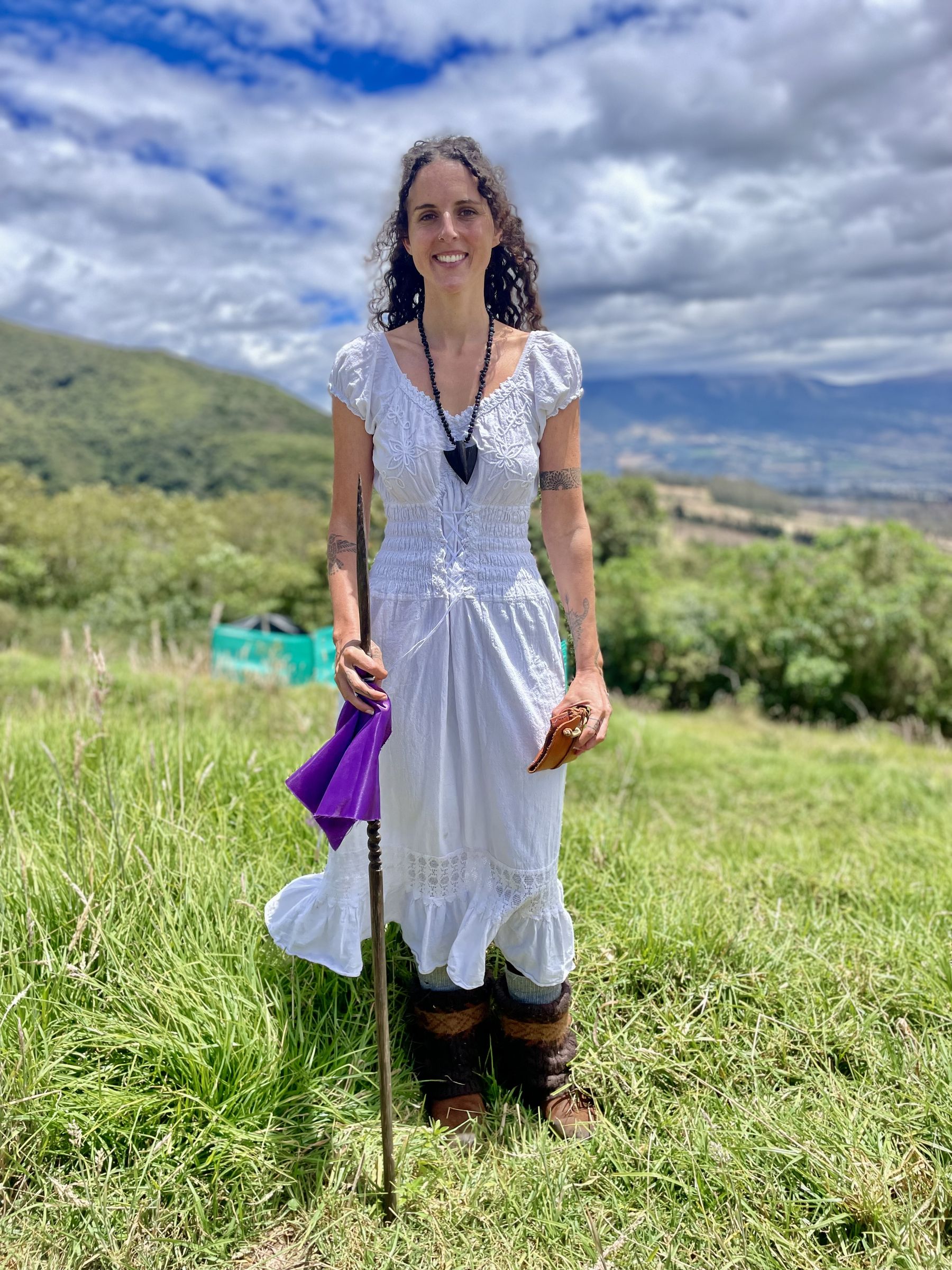 Alora standing in a mountain field in Ecuador holding a ceremonial staff with a purple prayer flag